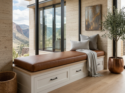 A modern built-in bench with a brown leather cushion and white storage drawers, styled with gray pillows and a throw blanket in a stone-walled room with large windows overlooking a mountain landscape.